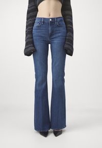 Sininen laajeneva farkut, joissa on korkea vyötärö, etutaskut ja tiukka ilme ylhäällä, levenevät halkiolla.