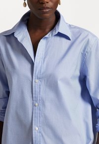 Donna che indossa una camicia leggera blu con bottoni e orecchini bianchi, labbra chiuse, su uno sfondo semplice.