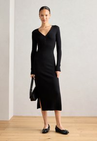 Abito midi a maglia a coste nere con maniche lunghe, scollo a V, dettaglio con bottoni e apertura laterale. Abbinato a scarpe flat nere e una piccola borsa a tracolla.