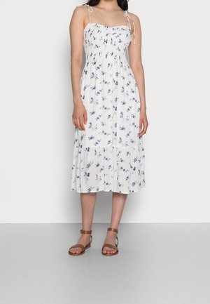 Femme portant une robe midi blanche à imprimé floral bleu avec des bretelles nouées aux épaules, associée à des sandales plates marron à bride cheville.