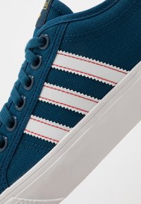 Modré plátěné tenisky s bílou gumovou podrážkou, námořnickými modrými tkaničkami a třemi bílými pruhy se zigzagovým okrajem a červeným prošíváním na boku.