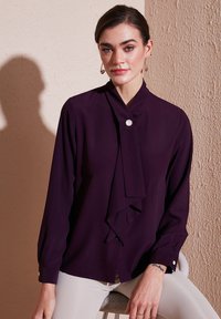 Blusa prugna con scollatura annodata, maniche lunghe e un unico bottone ai polsi. Tessuto morbido con una superficie liscia.