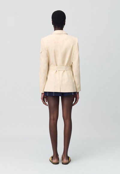 Blazer en lin beige avec une taille ceinturée, des fentes dans le dos et des épaulettes. Associé à une jupe bleue marine, mettant en avant un design minimaliste.