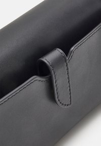Primo piano di un angolo di una borsa in pelle nera con cuciture visibili e un dettaglio di cinghia piegata su uno sfondo neutro.