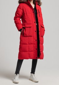 Lange rode gewatteerde jas met een afneembare bontkraag, voorzien van voorzakken, een rits aan de voorkant en drukknoopjes. Draagt over een zwarte top en broek.