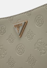 Surface en cuir beige embossée de motifs circulaires et d'étoiles, avec un logo triangulaire Guess USA doré et blanc comportant un point d'interrogation.