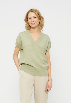 Kvinde med skulderlangt blondt hår iført en lysegrøn kortærmet V-hals sweater og beige bukser mod en ensfarvet baggrund.