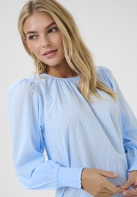 Blouse bleu clair avec des pois texturés, des manches ballon et des poignets smockés. Tissu doux avec un col rond.