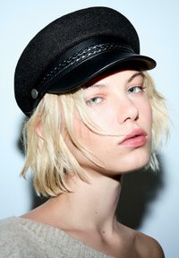 Ung person med kort blont hår som bär en svart vävd keps med läderkanter och en beige off-shoulder-tröja mot en ljus bakgrund.