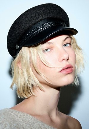 Młoda osoba z krótkimi blond włosami, nosząca czarną, plecioną czapkę z skórzanym wykończeniem oraz beżowy sweter bez ramiączek na jasnym tle.