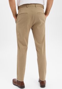 Beige jurkbroeken met een gladde textuur, smalle pasvorm, taps toelopende pijpen en twee achterzakken. Gecombineerd met bruine leren schoenen.