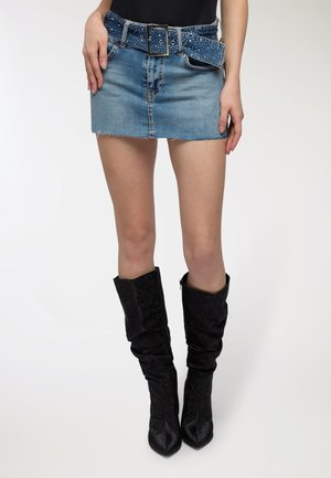 Gonna mini in denim con orlo sfrangiato, che presenta una cintura blu con borchie. Il modello indossa stivali neri con tacco a metà coscia di tessuto strutturato.