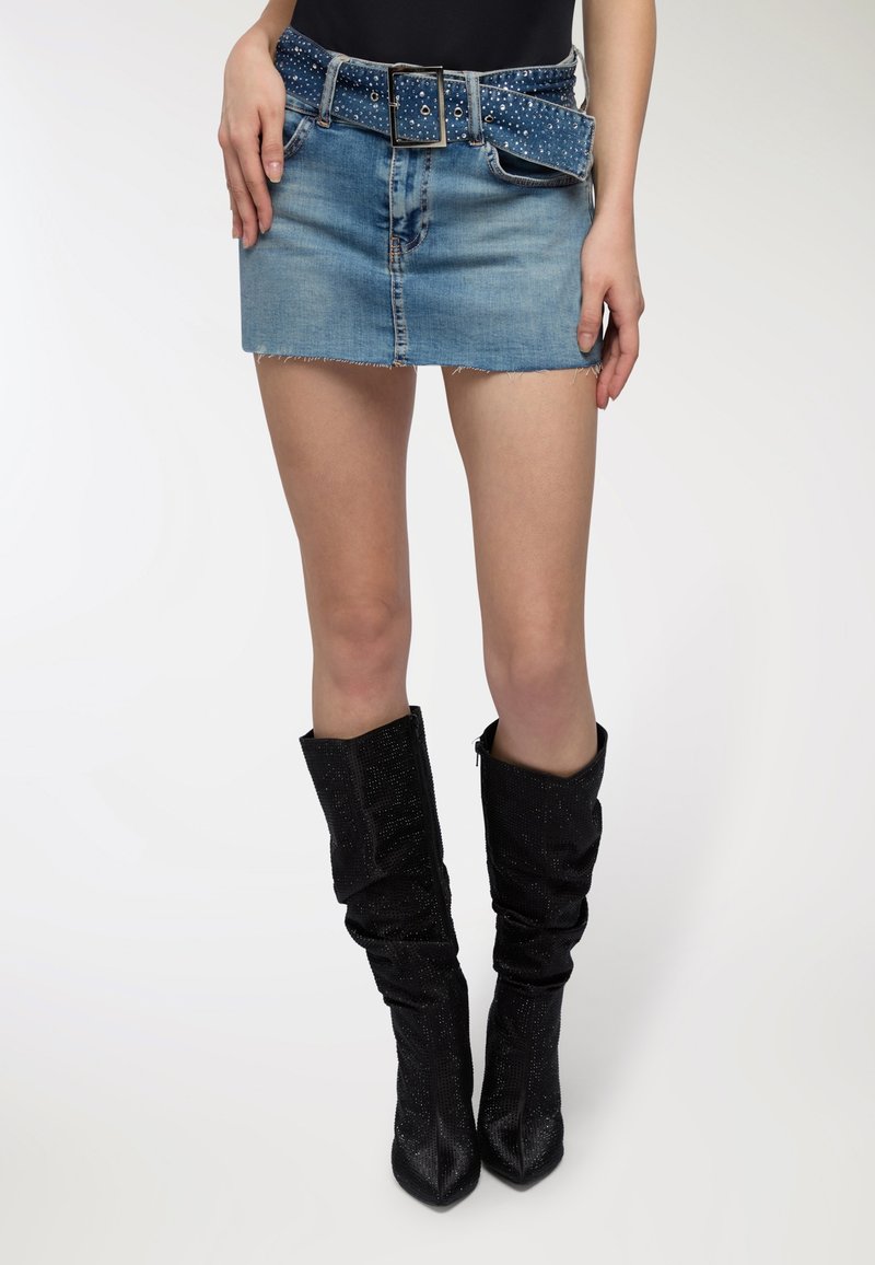 Gonna mini in denim con orlo sfrangiato, che presenta una cintura blu con borchie. Il modello indossa stivali neri con tacco a metà coscia di tessuto strutturato.