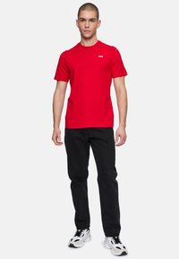 Rød kortærmet t-shirt med hvidt logo, parret med sorte jeans og hvide sneakers. Stoffet ser glat og afslappet ud.