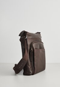 Brun læder skuldertaske med en tekstureret overflade, der har en frontlomme med lynlås og en justerbar strop. Firkantet form, minimalistisk design.