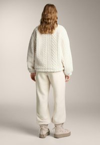 Fehér, bolyhos, rövidített kabát texturált kábelmintás kötött mintával, hozzáillő nadrággal párosítva. A bézs platform cipők kiegészítik az öltözéket.