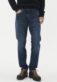 Vyras, dėvintis tamsiai mėlynus džinsus ir rudus batas su raišteliais, stovi su viena ranka kišenėje prieš baltą foną.