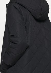 Chaqueta negra acolchada con capucha, tela texturizada y patrón geométrico en diamante. Detalles visibles en la costura lateral y en el bolsillo con cremallera.