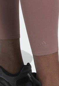 Leggings atletici rosa con una texture liscia, con un piccolo logo Adidas in rilievo vicino all'orlo. Indossati con scarpe da ginnastica nere.