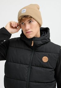Svart polstret vest med stor quilt, mørke ermer og en beige strikket lue med etikett. Lærpatch-detaljer og justerbar hette.