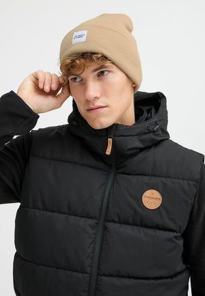 Black padded vest with large quilting, dark sleeves, and a beige knit beanie with a label. Leather patch accents and adjustable hood.