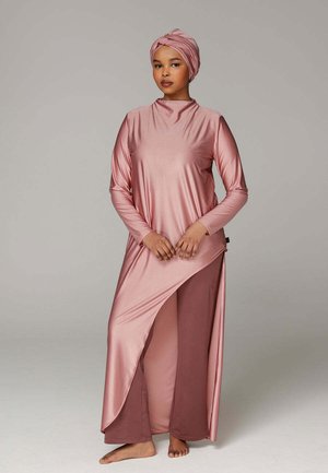 Femme portant un ensemble tunique et pantalon rose brillant avec un foulard assorti sur la tête, debout pieds nus devant un fond gris uni.
