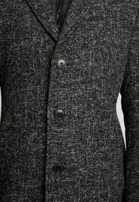 Manteau gris texturé avec une silhouette ajustée, comportant trois boutons noirs et un col à revers. Doublure intérieure visible.