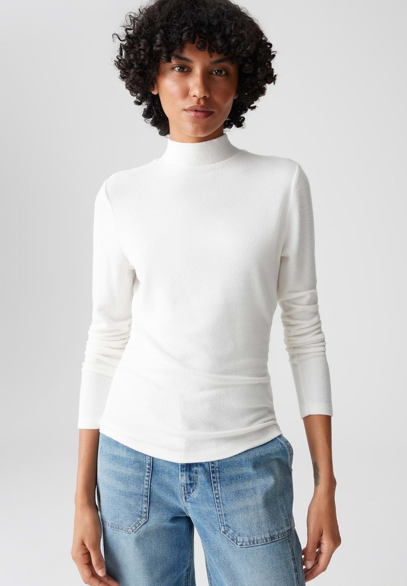 Weißes Langarm-Turtleneck-Oberteil aus einem weichen, elastischen Stoff mit figurbetontem Schnitt, kombiniert mit hellblauen Denim-Jeans mit aufgesetzten Taschen.