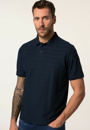 Marineblauwe polo met subtiele horizontale strepen, korte mouwen, een kraag en een knoopsluiting met één knop. Draagt bij denim jeans.