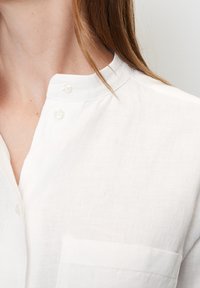 Weißes Leinenhemd mit Stehkragen, kleinem Brusttasche und Knopfdetails nahe dem Kragen. Glatte Textur, leichter Stoff.