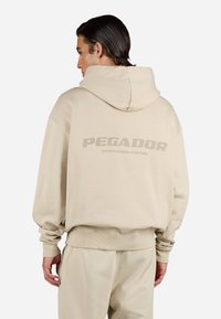 Beige hettegensere med en stor "PEGADOR" logo på baksiden. Laget av mykt materiale, har en romslig hette og ribbestrikkede mansjetter.