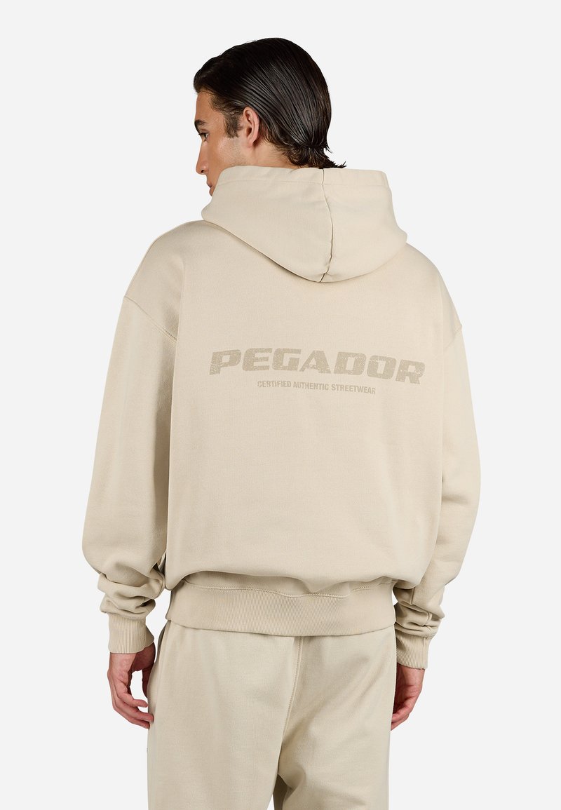 Beige hettegensere med en stor "PEGADOR" logo på baksiden. Laget av mykt materiale, har en romslig hette og ribbestrikkede mansjetter.
