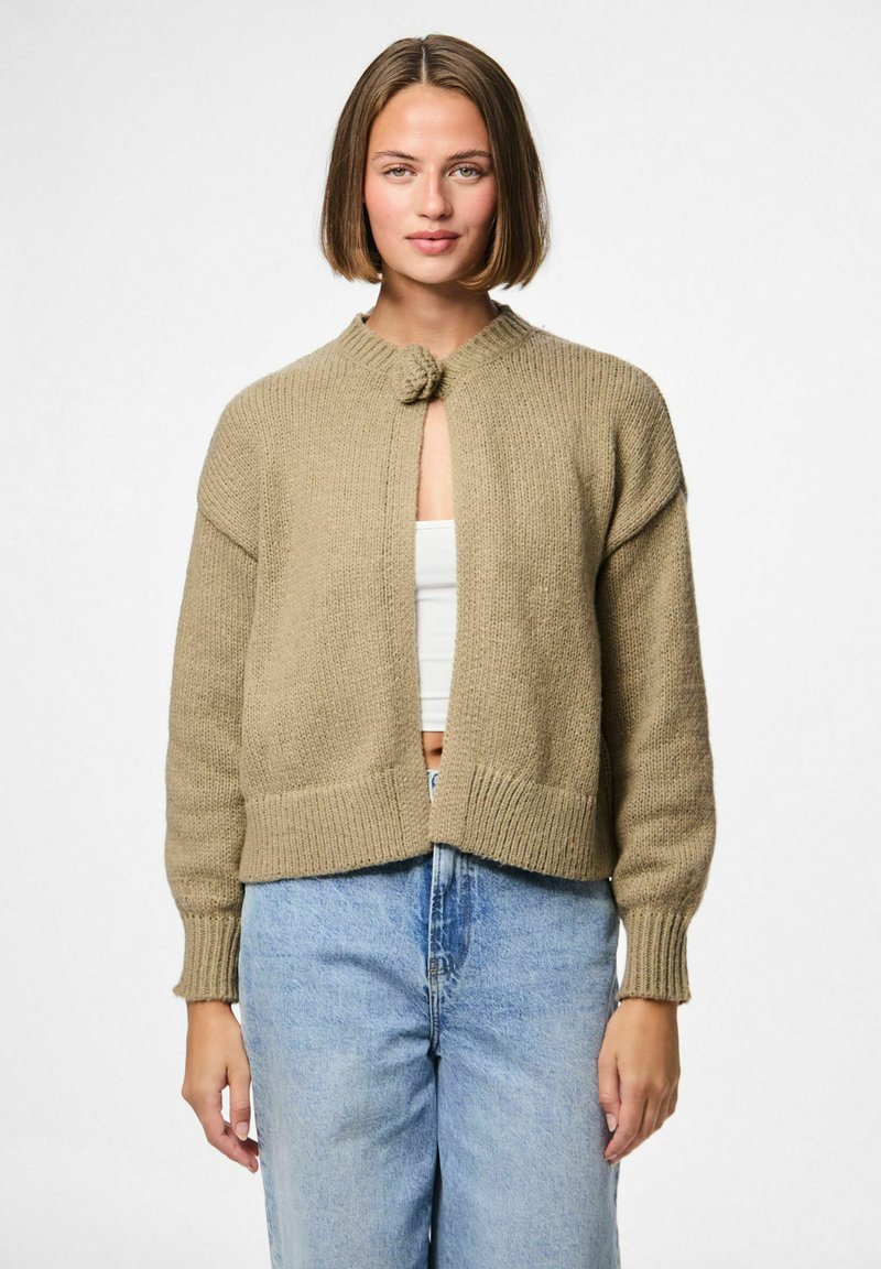 Beige knitted cardigan with a round collar, front closure featuring a decorative knot, and ribbed cuffs, worn over a white top and blue jeans.