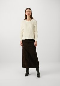 Off-white strikkegenser med tekstur, kombinert med en brun denim maxiskjørt. Modellen har på seg svarte heler. Enkelt, uformelt utseende.