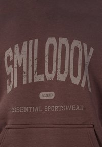 Sudadera marrón con el texto desgastado "SMILODOX 2013© ESSENTIAL SPORTSWEAR" y un bolsillo frontal visible en la parte inferior.