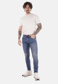 Uomo con capelli ricci e tatuaggi su entrambe le braccia che indossa una t-shirt bianca, jeans blu e sneakers bianche, in piedi con una mano in tasca.