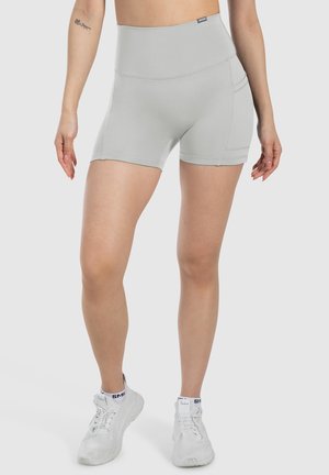 Kvinde iført lysegrå atletikshorts med høj talje og sidelomme og hvide atletikskoninger mod grå baggrund.