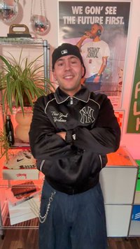 Jauns vīrietis melnā Ņujorkas "Yankees" jakā un cepurē stāv ar savelkšanas rokām, smaidot telpā, kurā ir augi, disko bumbiņas un motivējošs plakāts.