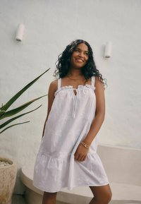 Witte katoenen jurk met een rimpelronding en strik, voorzien van geborduurde palmbomenpatronen. Losse pasvorm en gelaagd rokdesign.