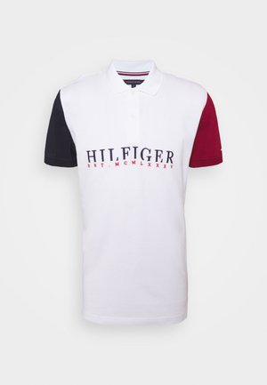 Biała koszulka polo z granatowym lewym rękawem i czerwonym prawym rękawem, logo "HILFIGER" oraz rzymskie cyfry w kolorach granatowym i czerwonym na wysokości klatki piersiowej.