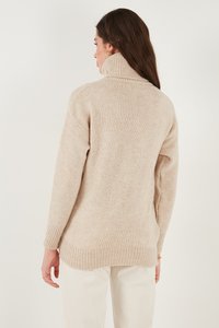 Pulover bej cu guler înalt, din tricot texturat, cu mâneci lungi și manșete și tiv din bumbac. Purtați cu pantaloni de culoare deschisă, văzut din spate.