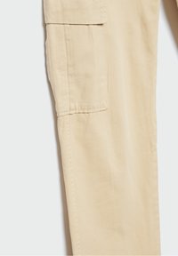 Beżowa nogawka spodni z prostokątną kieszenią cargo z klapą i widocznym podwójnym przeszyciem wzdłuż szwów.