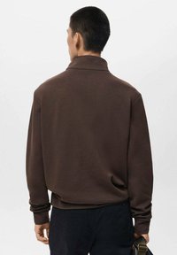 Pullover marrone a maniche lunghe con colletto, caratterizzato da una vestibilità morbida e una texture morbida. Indossato con pantaloni scuri, che mettono in evidenza la semplicità nel design.
