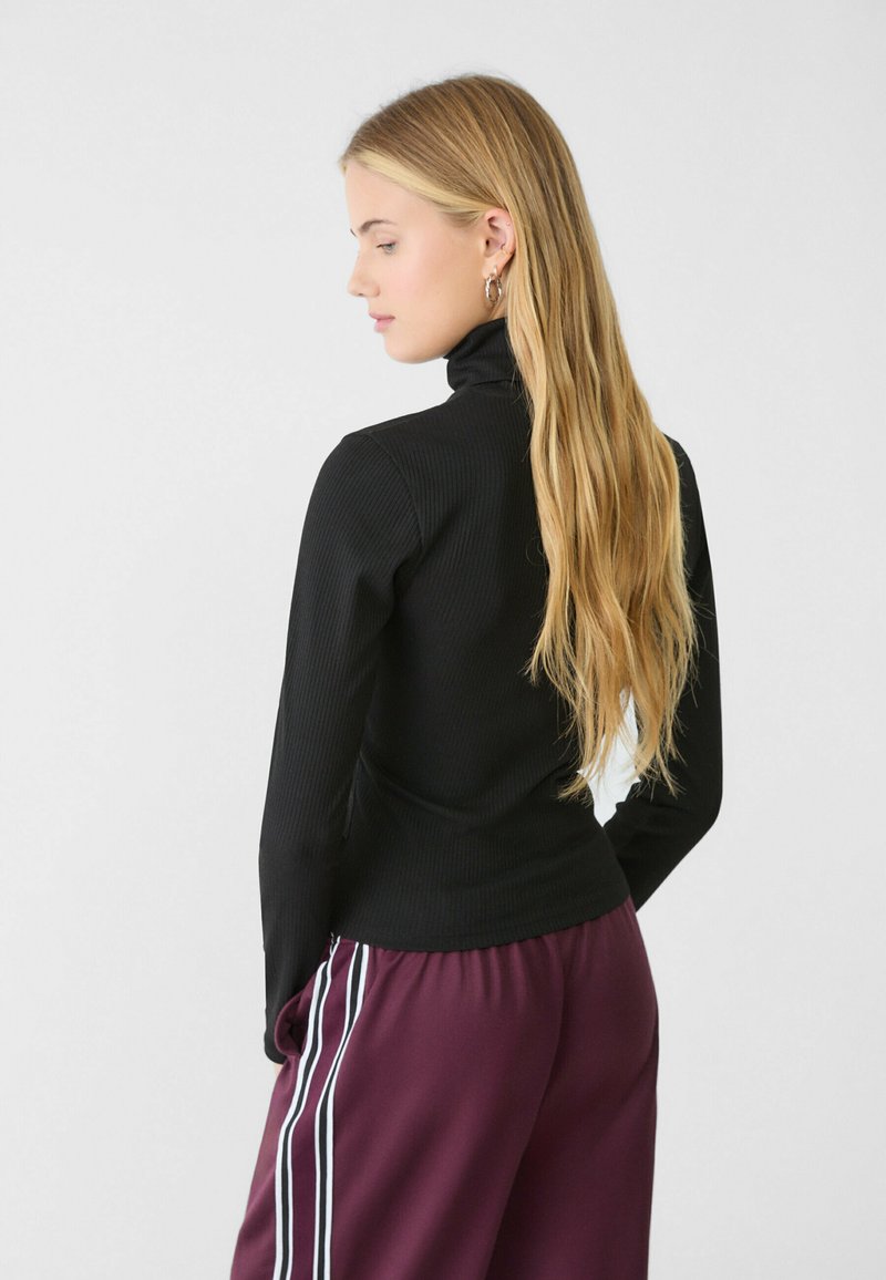 Top a collozzo nero a costine con maniche lunghe, abbinato a pantaloni bordeaux con strisce bianche ai lati. Texture liscia e aderente. Vista posteriore.