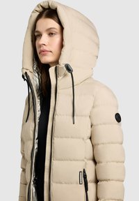 Doudoune beige avec une grande capuche, motif matelassé, accents de fermeture éclair noirs et une doublure intérieure brillante. Dispose de cordons de serrage à la capuche.