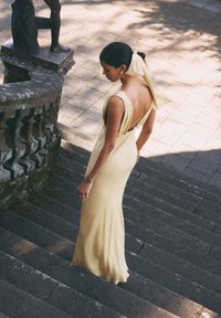 Robe longue et ajustée de couleur jaune avec un dos décolleté, des bretelles et un tissu fluide. Un mannequin descend des marches en pierre à côté d'une sculpture.