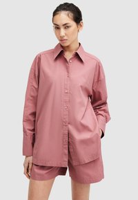 Camisa de botones rosa de gran tamaño con cuello, mangas largas y una textura suave. Combinada con shorts a juego, presenta un ajuste relajado y un diseño minimalista.