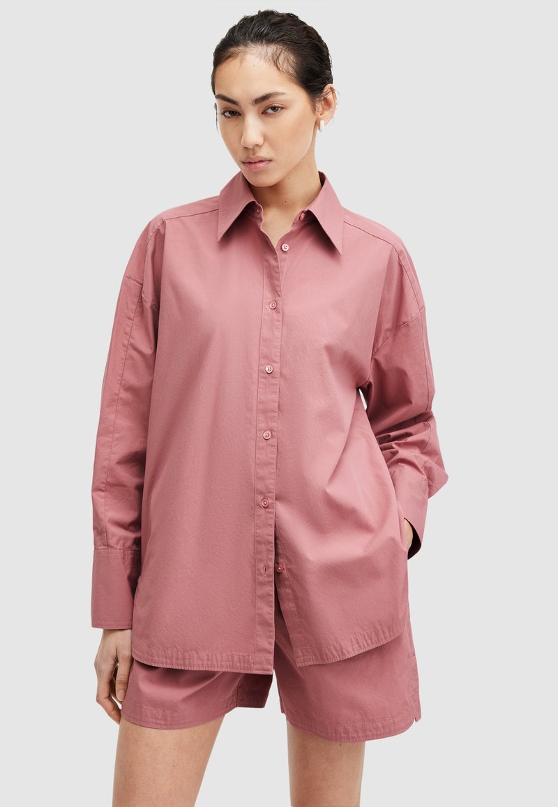 Camisa de botones rosa de gran tamaño con cuello, mangas largas y una textura suave. Combinada con shorts a juego, presenta un ajuste relajado y un diseño minimalista.