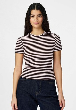 Młoda kobieta o długich ciemnych włosach, ubrana w krótki rękaw, czarno-białą, paskowaną bluzkę i ciemnoniebieskie dżinsy, stojąca na tle białego tła.