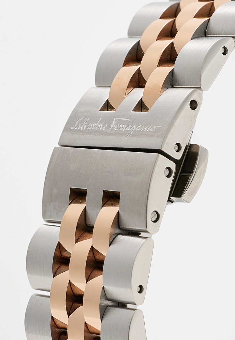 Reloj de pulsera metálico con una pulsera de acero inoxidable y tono rosa, que presenta una textura cepillada, eslabones entrelazados y cierre de hebilla.